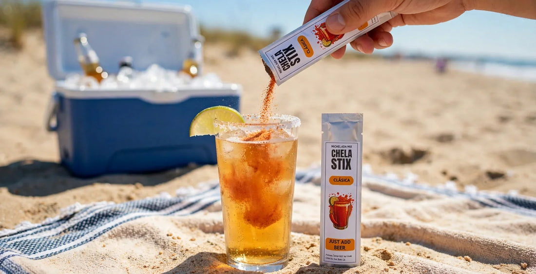 A hand pours a Chela Stix Clásica michelada mix stick into a tall glass of beer over ice on a beach towel, with a second stick standing upright beside the glass and a blue cooler full of beer bottles on the sand behind it, bright sunny beach day.