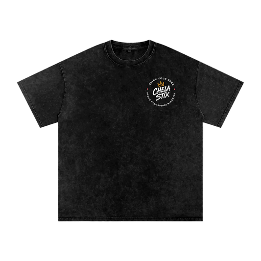 A black acid-wash short-sleeve t-shirt with a circular white graphic on the left chest reading 'CHELA STIX' and 'MAKE YOUR MARK'. The t-shirt is displayed flat against a solid white background.