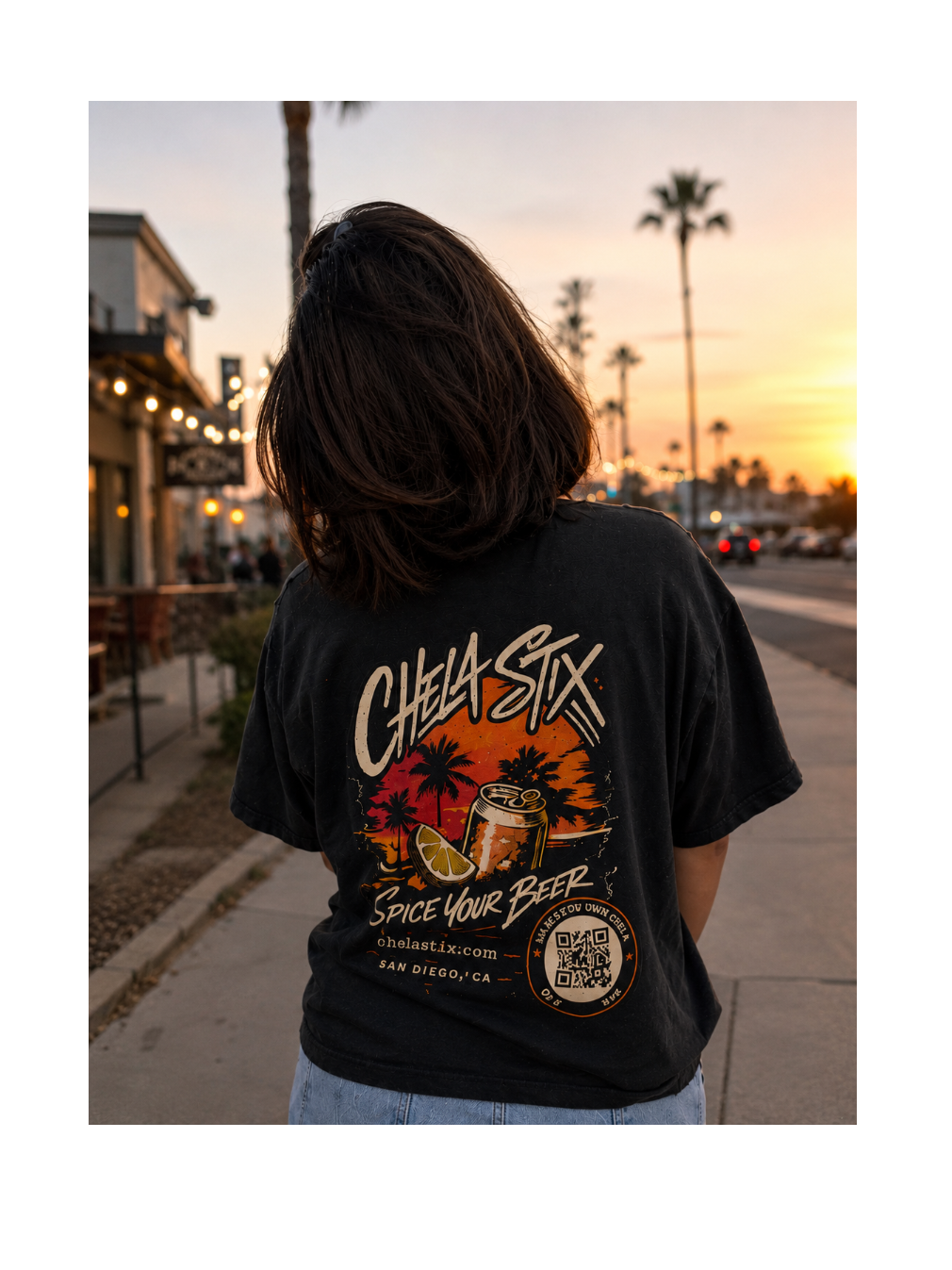 A person viewed from behind wearing a black graphic t-shirt with a colorful 'Chela Six' beer design on the back, standing on a sidewalk during sunset.