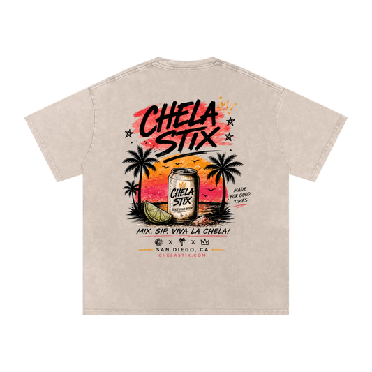 A light beige graphic t-shirt shown from the back, featuring a colorful sunset illustration with palm trees, a beer can labeled 'Chela Stix', and bold black text.